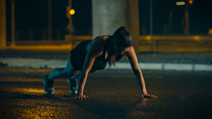 Beautiful Energetic Fitness Girl in Black Athletic Top and Shorts is Doing Push Up Exercises. She is Doing a Workout in an Evening Wet Urban Environment Under a Bridge.
