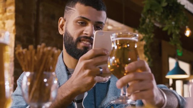 Cheerful Arabian man taking a photo of glass with light beer at the table in bar