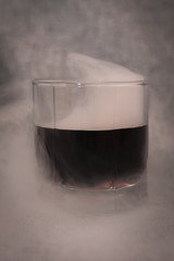 glass of dark liquid and smoke around on a gray background