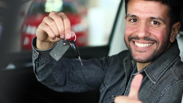 Positive young man holding a key sitting in a car with thumb up