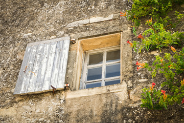 Very old and weathered window with shutter surrounded by orange trumpet flowers in the Mediterranean masonry facade of an old French house.