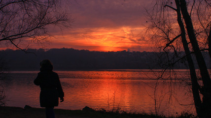 Naklejka na ściany i meble Silhouette of a woman walking in front of a lake at sunset in winter with dramatic sky