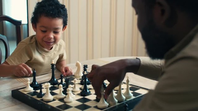 Panning shot of African American boy trying to play chess with his dad at home