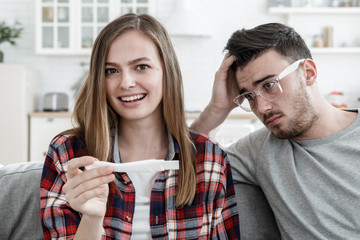 Young lady glad that she has got pregnant and her boyfriend looking worried and frightened with puzzled face not ready for family relationship