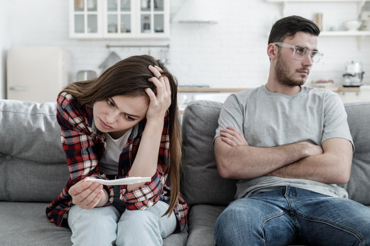 Young man and woman sitting in living room looking depressed because of pregnancy of lady, not knowing what to do