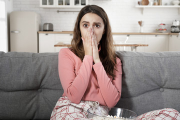 European lady watching TV and looking frightened with round eyes, having seen something shocking on screen