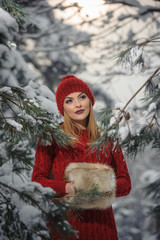 Beautiful woman in red with brown fur cape enjoying the winter scenery in forest. Blonde girl posing under snow-covered trees branches. Young female with snowflakes around in bright cold day, makeup