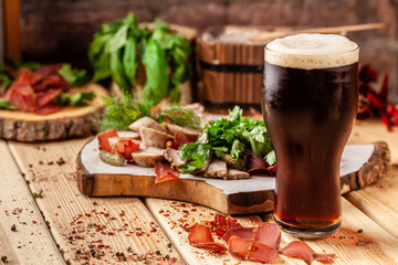 Concept of Georgian restaurant. A glass of black beer with foam stands on a wooden table, next to it is a beer snack, dried sausage, basturma, smoked meat and parsley with dill.
