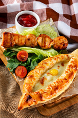 Traditional georgian adzharian khachapuri and meat shashlik with vegetables at decorated wooden board background.