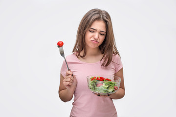 Bored young woman look at glass bowl with salad. She hold tomato on fork in another hand. Model is...