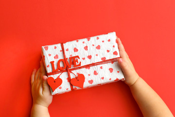 Child hands holding beautiful gift box on red background. Holiday, bright and festive view, flat lay. Valentines day greeting banner. Father day, Mother day or Birthday celebration.