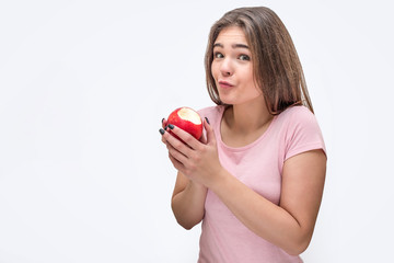 Nice and attractive young woman look on camera and hold bitten apple. She is surprised. Isolated on white background.