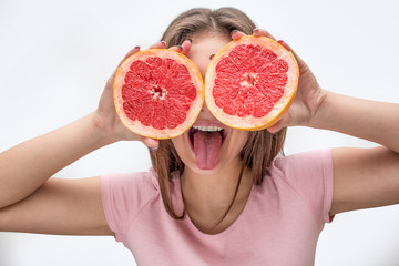Playful young woman cover eyes with two halfs of grapefruit. Model show tongue. Isolated on grey background.
