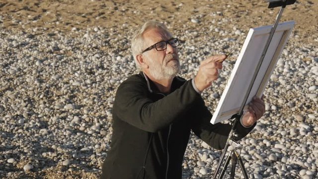 Senior man paints a picture on the beach. High angle view of elderly male artist painting abstract picture with a brush at pebble sunrise sea beach.