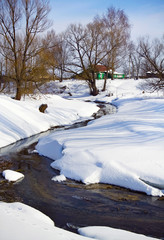 Winter rural landscape with the small river and a rural lodge against the background of. The small river passes through all image on diagonal. Clear day. Shadows of trees beautifully lay down on snow.