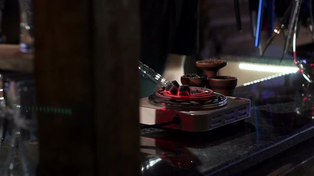 Kindling of square coal for a hookah on a special furnace with a hot spiral. Close-up of kindling coals for hookah in hookah bar. Man holds red-hot coal with tongs.