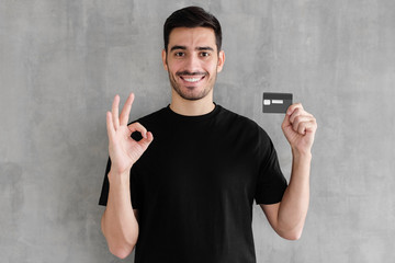 Young man in black t-shirt, holding credit card and showing OK sign, standing against gray textured wall
