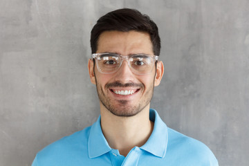 Smiling modern nice guy in blue polo shirt and trendy trasparent eyeglasses, standing against gray textured wall