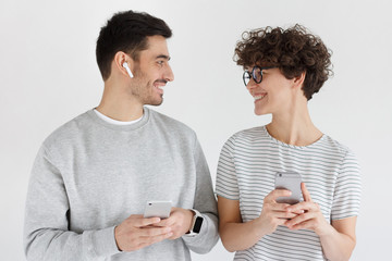 Young happy couple holding smartphones, standing together, looking at each other, isolated on gray background