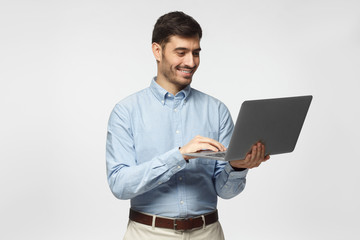 Young handsome business man standing, holding laptop with happy smile, typing, using touchpad