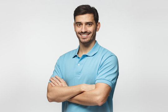 Portrait of attractive young sporty man in blue polo shirt standing with crossed arms, isolated on gray background