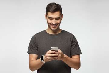Young european male standing isolated on gray background, looking at screen of phone with smile,...