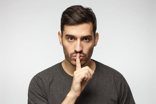 Young man dressed in casual t-shirt, pressing finger to lips as if asking to keep silence,  isolated on gray background. Shhh gesture