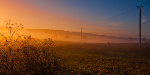 Fog on the countryside, next to the poles with wires, orange glowing fog, bright colors,
