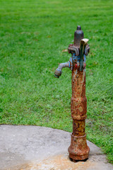 A portrait of a vertical and old fashioned rustic water fountain made of iron, with many rust points, over a concrete base and a green grass background gradually out of focus