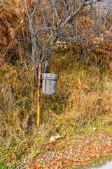 A wooden rubbish bin on a path in the forest. In front of a bare tree leaves, and on the ground yellow and brown leaves due to autumn
