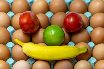 A funny artistic face made of a tray with eggs as the background, two tomatoes as the eyes, one lemon as the nose and a banana as the mouth