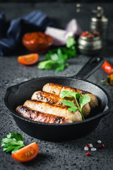 Grilled sausages in cast iron skillet with herbs, sauce and spices on dark background. Selective focus, space for text.