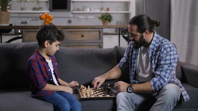 Handsome bearded father teaching mindful little mixed race son chess while sitting on sofa in domestic room. Caring stay-at-home dad developing intellectual skills of his boy with chess game at home.