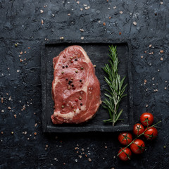 Raw beef rib eye steak with tomatoes and rosemary on black background top view. Square crop.