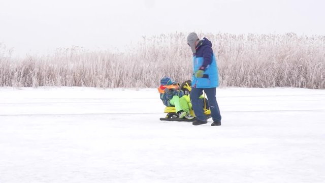 dad in blue ski suit and three happy children in warm winter clothes ride on yellow sled on frozen snow-covered river.  Concept of friendly sports family. Winter holidays in village. Christmas eve