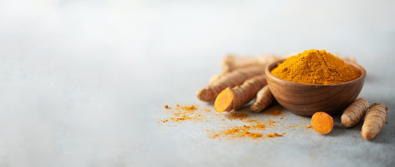 Turmeric powder in wooden bowl and fresh turmeric root on grey concrete background. Banner with...