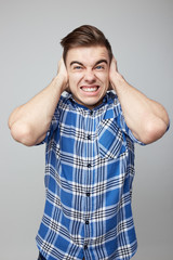 Scared guy dressed in a plaid shirt and jeans closes his ears with his hands on a white background in the studio