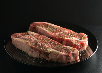 Two raw strip steaks on black plate with seasoning, side angled view