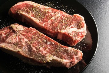 Two raw strip steaks on glossy black plate with seasoning. Angled top view.