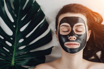 Attractive young  girl with eyes closed lying on bed relaxing with applied a black mask to her face  with a green leaf enjoying and relaxing , day spa . Skin care and beauty concept.