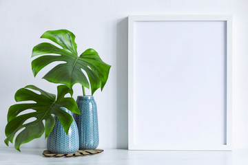 Modern and minimalistic composition of mock up photo frame with green tropical leafs in design vase. Stylish concept of mockup frame. White backgrounds wall. 