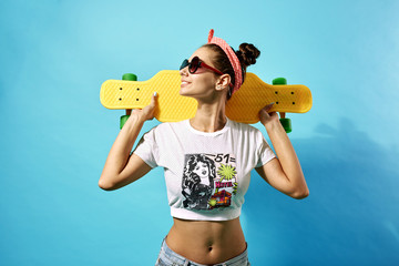 Funny young girl in sunglasses and pink bow on her head dressed in jeans and top stands with yellow skateboard on the back on the blue background in the studio