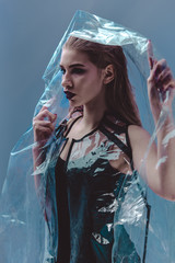 Fashionable model with wet hair dressed in black swimming suit and transparent rain coat is posing under the transparent membrane in the studio with lighting simulating evening street light