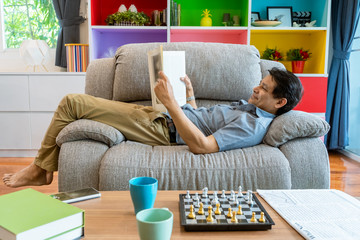 Obraz premium Asian middle-aged man lying on the sofa reading a magazine, very comfortable