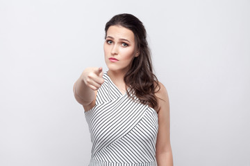 You. Portrait of serious beautiful young brunette woman with makeup and striped dress standing, looking and pointing at camera. indoor studio shot, isolated on grey background.