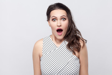 Portrait of amazed beautiful young brunette woman with makeup and striped dress standing, looking at camera with big eyes, open mouth and shocked face. indoor studio shot, isolated on grey background.