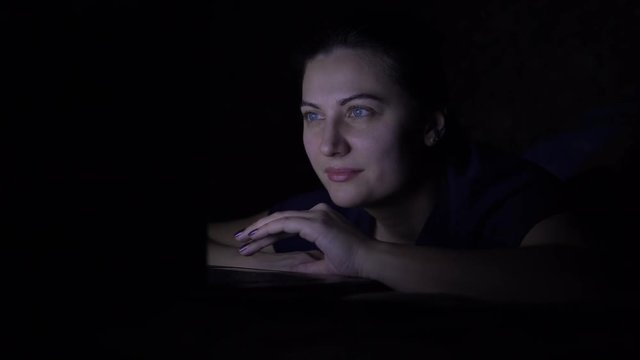 A woman works using a mobile computer while lying on the bed at home at night. Isolated light