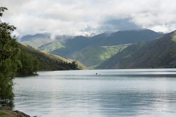 Fototapeta premium A rowing scull on Lake Dustan on a calm, but cloudy day, surrounded by snow topped hills. Central Otago, New Zealand.