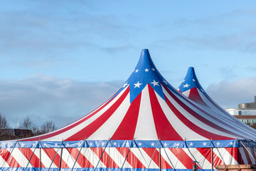 Red and white circus tent topped with bleu starred cover against a sunny blue sky with clouds