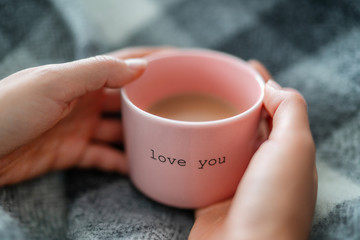 Winter warm coffee cup woman relaxing at home cozy with wool blanket - morning breakfast girl drinking with the words LOVE YOU written on pink mug for valentines day. Romantic quote.